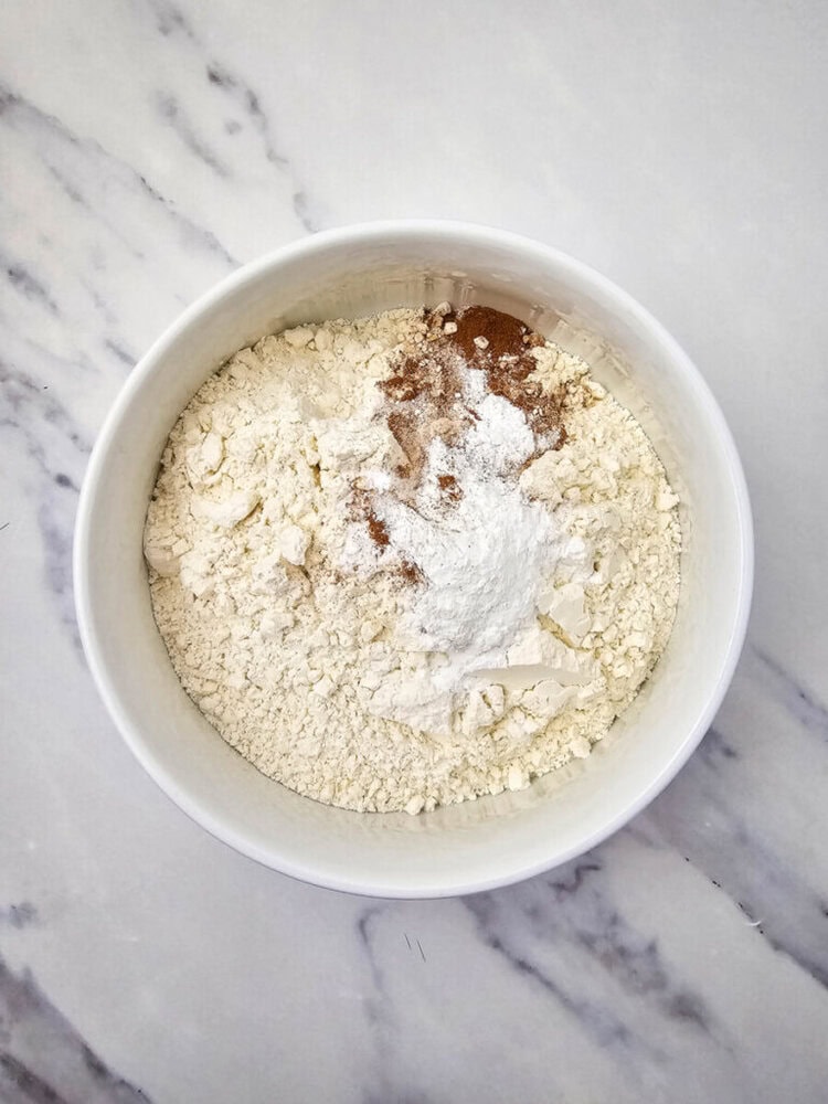 A white bowl on a marble countertop containing flour, baking powder, and a sprinkle of cinnamon.