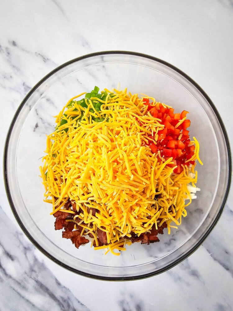 Glass bowl with ingredients including shredded cheese, diced red and green bell peppers, and ground meat on a marble surface.