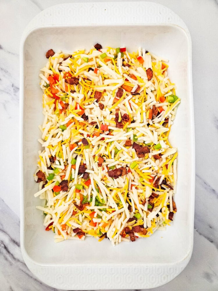 A white baking dish filled with shredded cheese, chopped bacon, and colorful vegetable pieces on a marble surface.