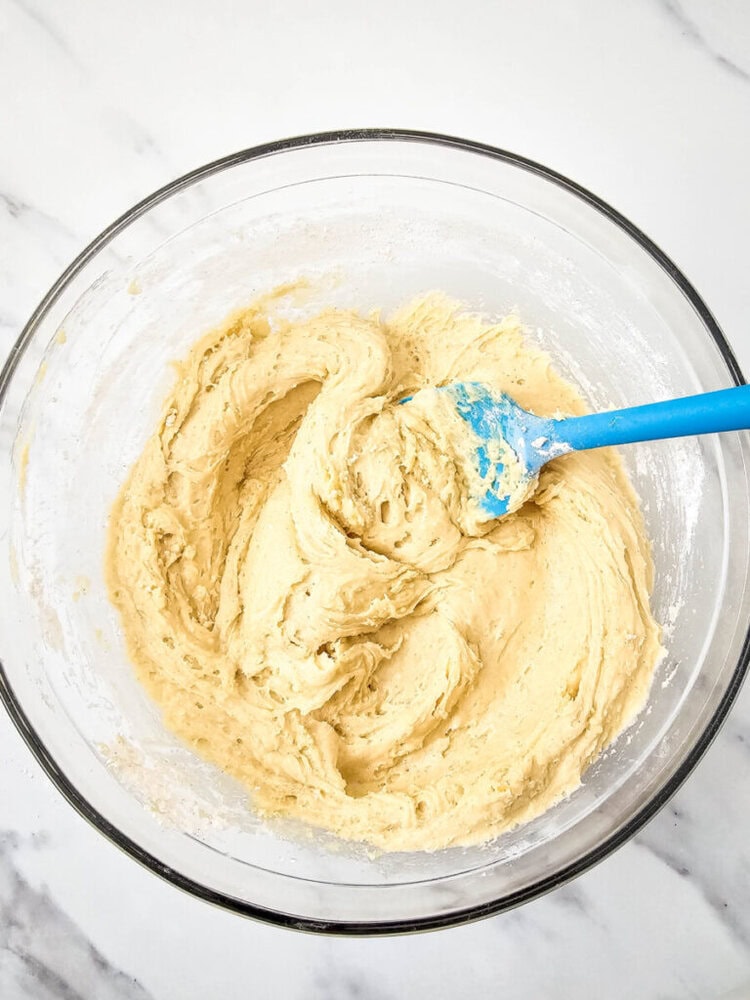 A bowl of creamy cake batter is being mixed with a blue spatula on a white marble surface.