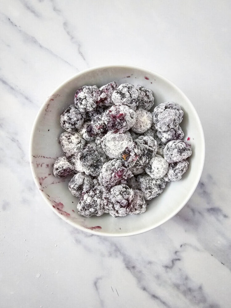 A bowl of blackberries coated in a layer of sugar on a marble surface.