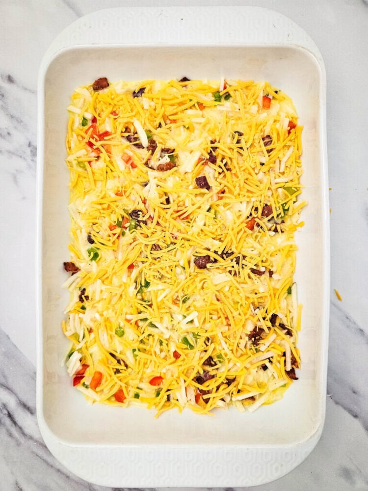A white baking dish filled with an uncooked layered casserole. It contains shredded cheese, ground meat, green and red bell peppers, over a creamy base. The dish is on a marble surface.