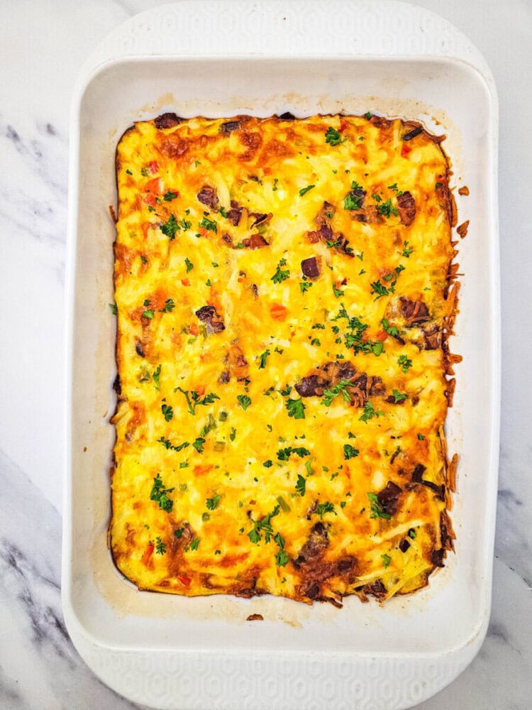 Baked casserole with melted cheese, diced meat, and chopped parsley in a white rectangular dish on a marble surface.
