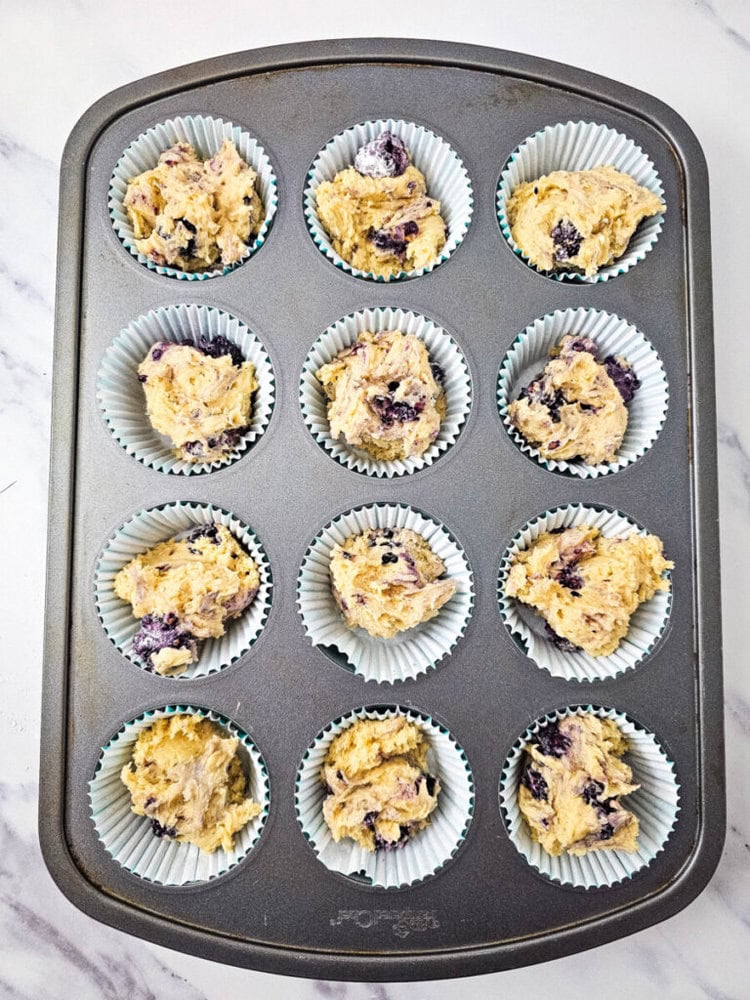 Muffin tin with 12 paper liners, each filled with uncooked blueberry batter, on a marble countertop.