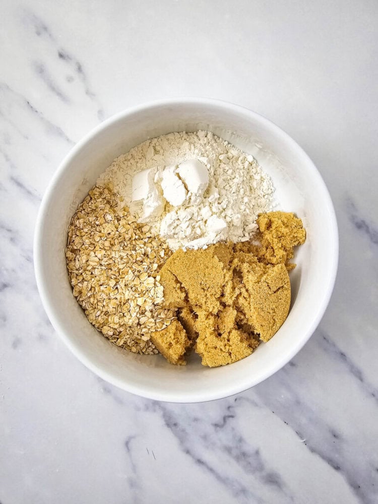 A white bowl containing rolled oats, flour, and brown sugar, arranged separately on a marble surface.