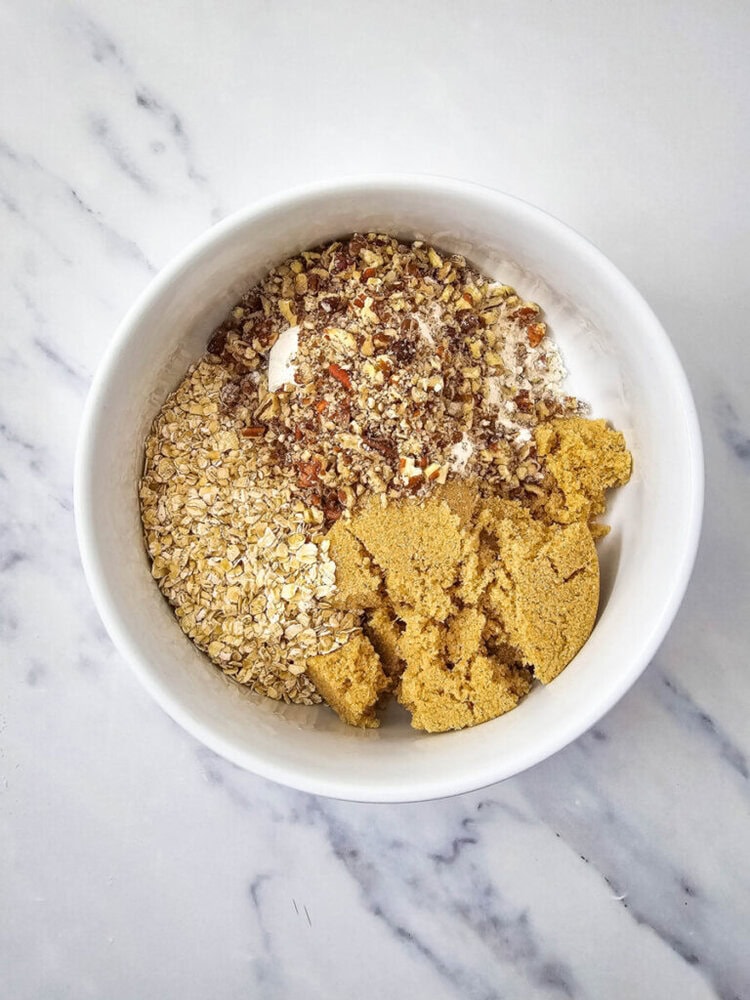 A white bowl containing oats, brown sugar, and chopped nuts on a marble surface.