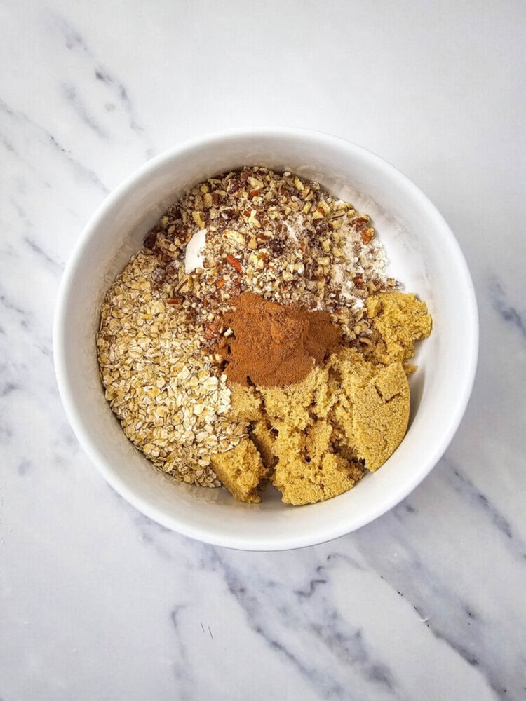 White bowl containing oats, chopped nuts, brown sugar, and cinnamon on a marble countertop.