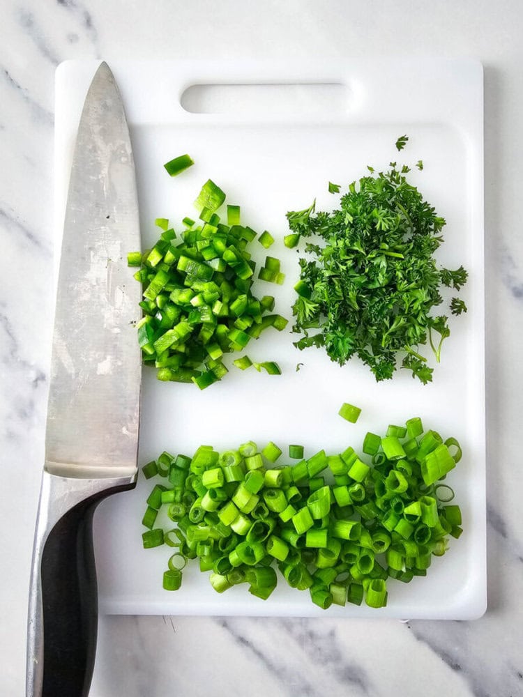 A cutting board with chopped green onions, parsley, and green chili. A knife is placed on the left side of the board.