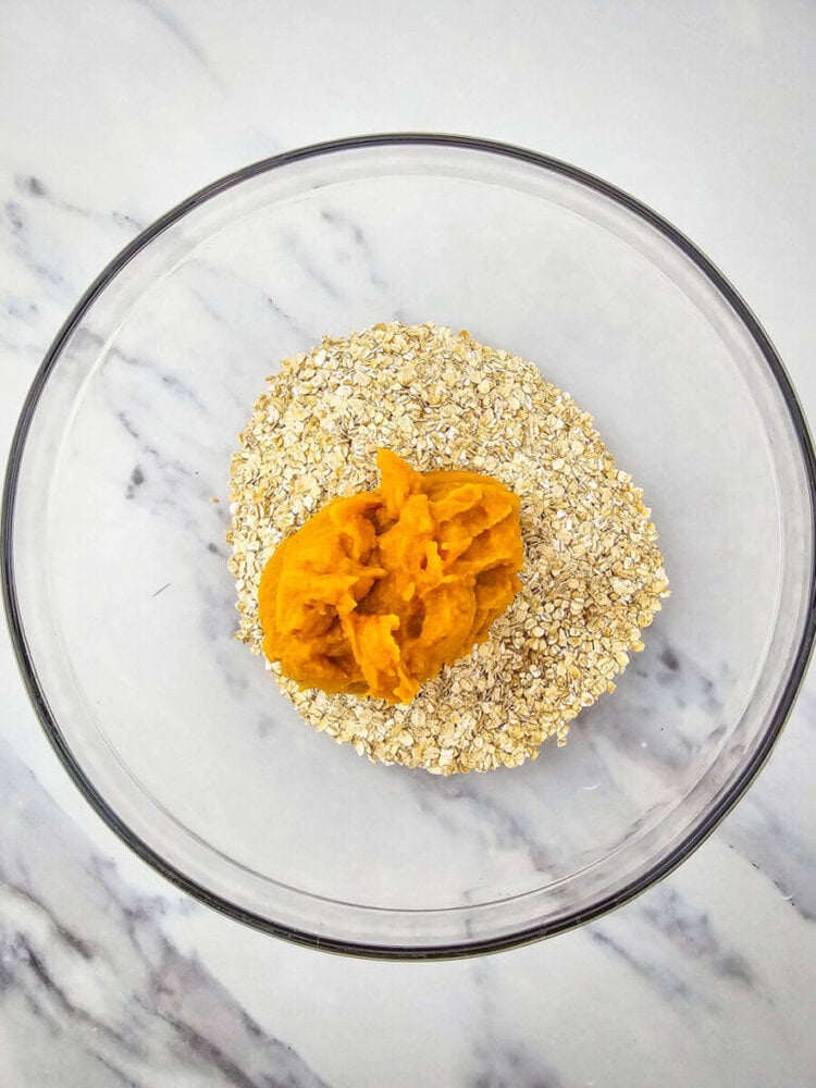 A glass bowl containing oats and a dollop of mashed sweet potato on a marble countertop.