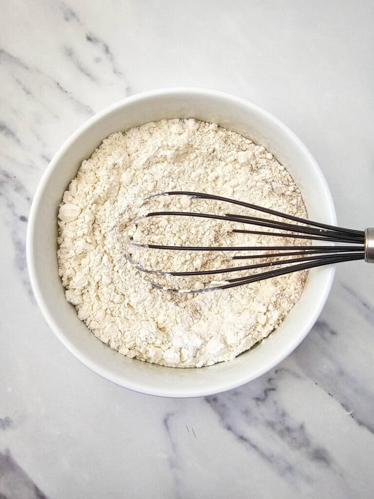 A whisk resting in a white bowl containing flour, placed on a marble surface.