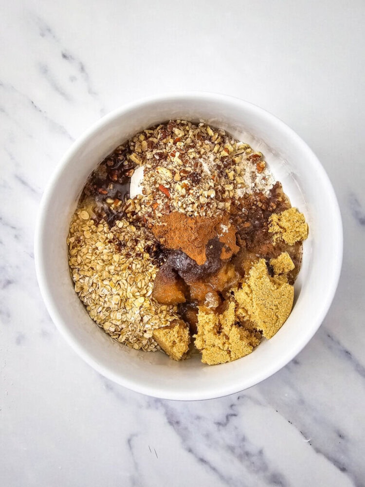 A white bowl containing oats, brown sugar, cinnamon, ground flaxseed, and a dark syrup on a marble surface.