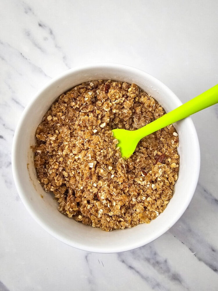 A white bowl filled with a mixture of oats, nuts, and brown sugar, with a green spatula resting inside, placed on a marble surface.