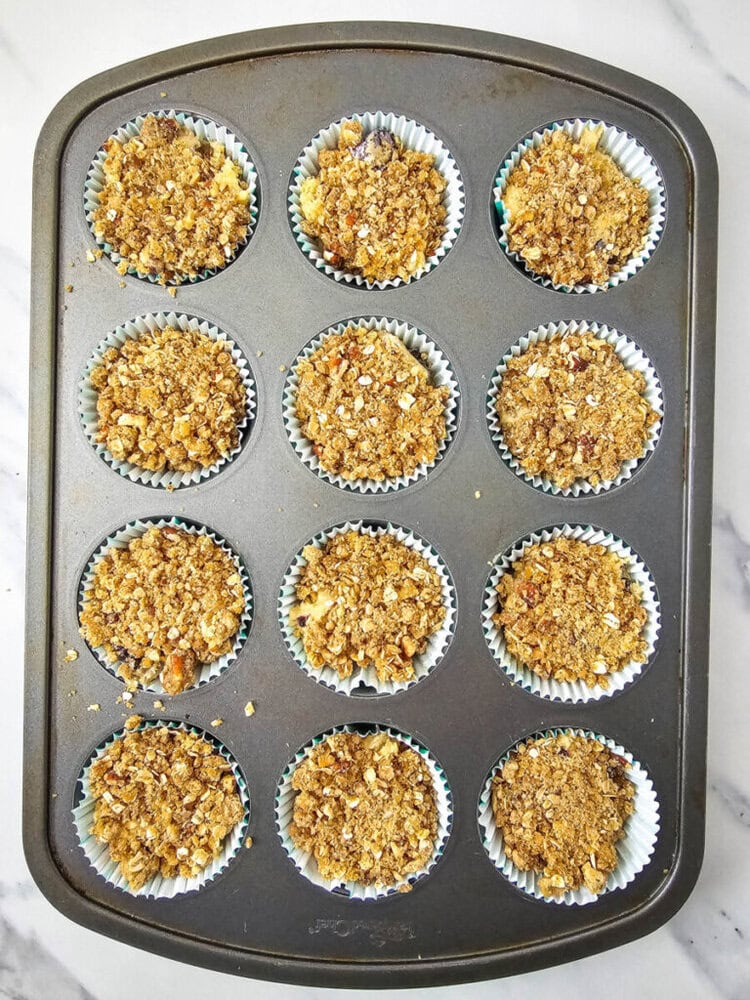 Muffin tray with twelve unbaked crumb-topped muffins on a marble surface.
