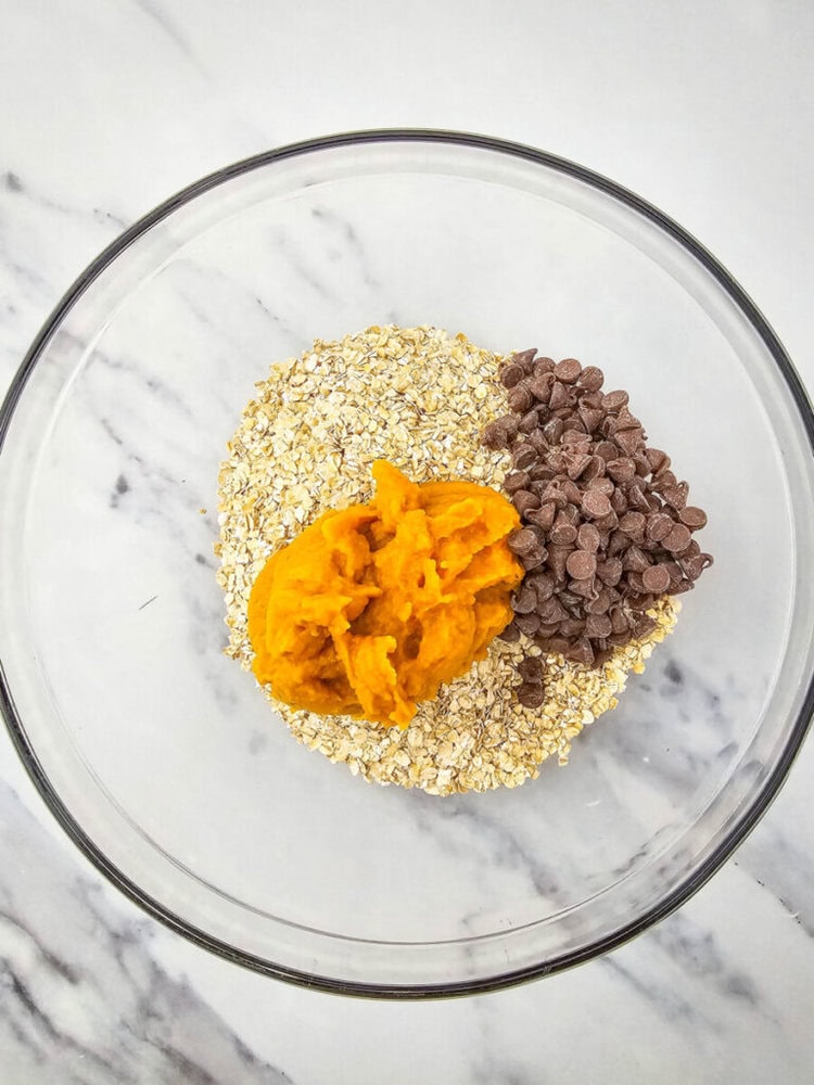 A glass bowl contains oats, pumpkin puree, and chocolate chips on a marble surface.