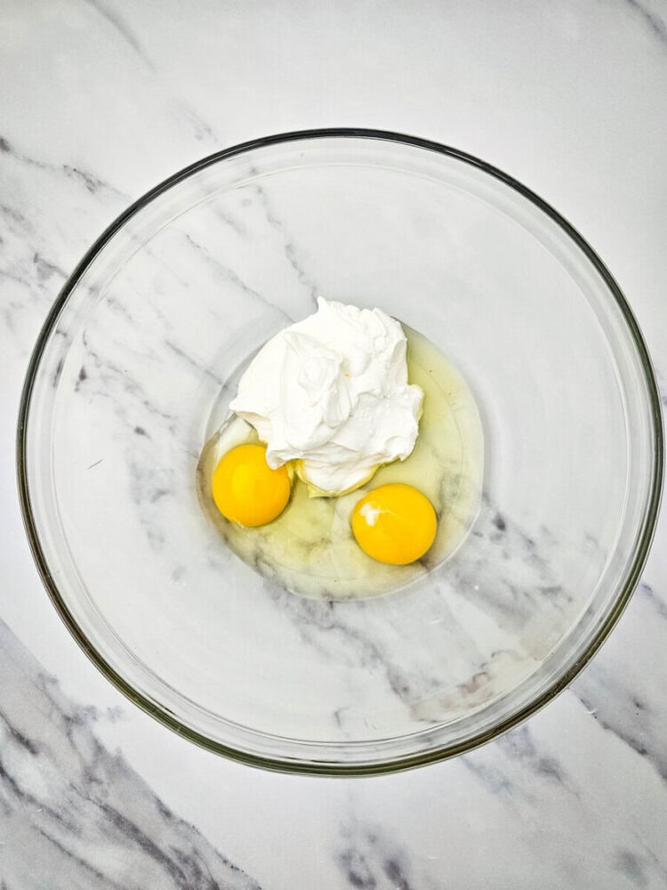A clear glass bowl contains two raw eggs and a dollop of white cream on a marble surface.