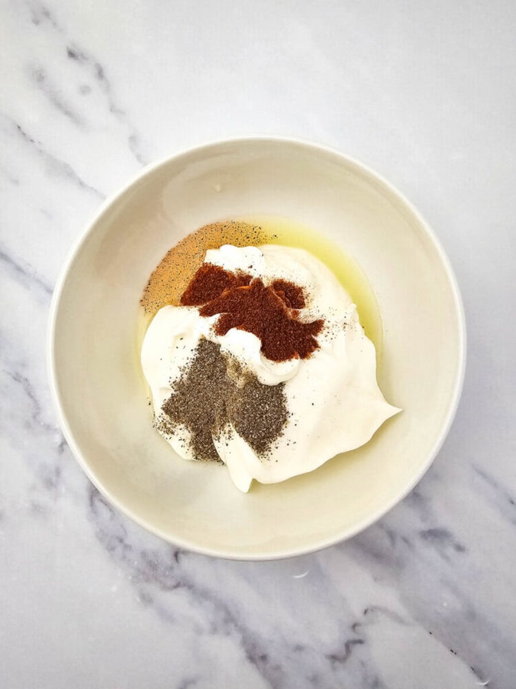 A white bowl contains mayonnaise, garlic powder, and black pepper, placed on a marble surface.