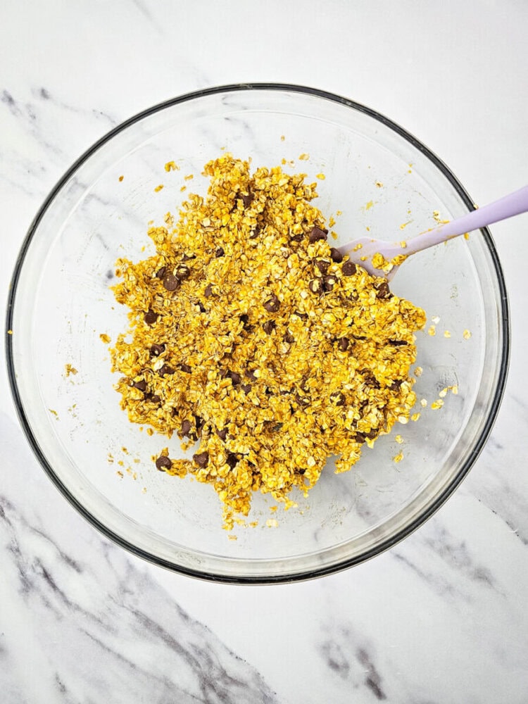 A glass bowl filled with a mixture of oats, chocolate chips, and a yellow ingredient, possibly pumpkin or turmeric, with a spoon resting in it, on a marble surface.