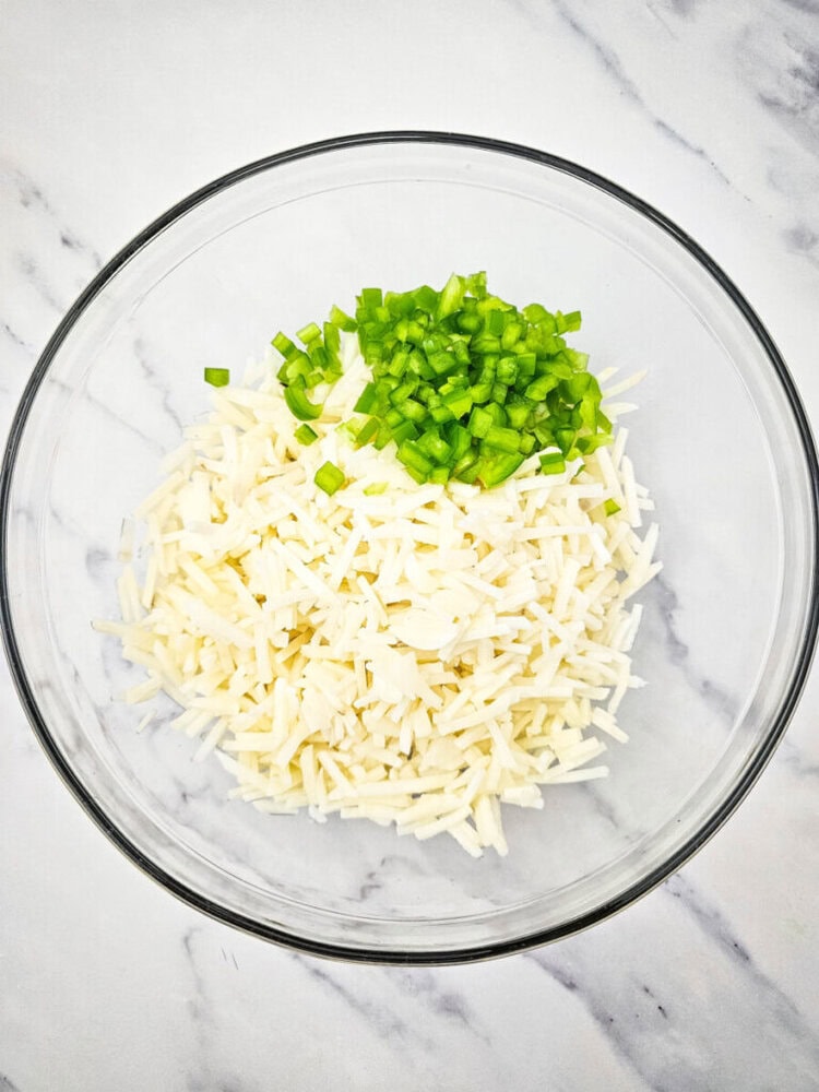 A glass bowl contains shredded potatoes and chopped green bell peppers on a white marble surface.