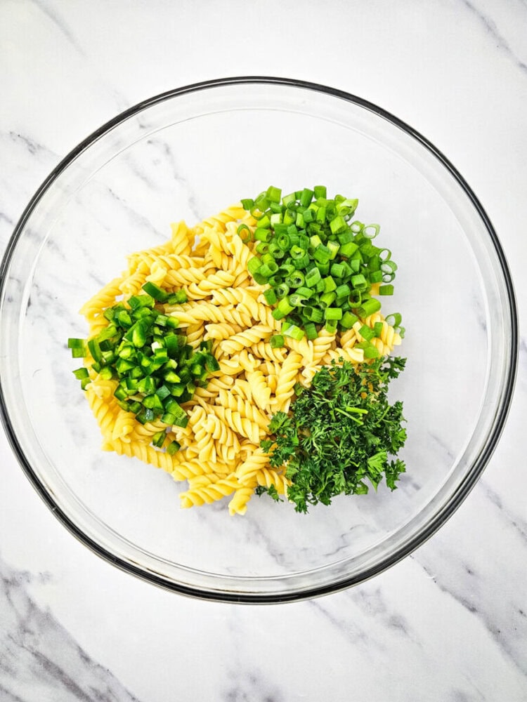 A bowl containing uncooked fusilli pasta, chopped green onions, diced green peppers, and parsley on a marble surface.