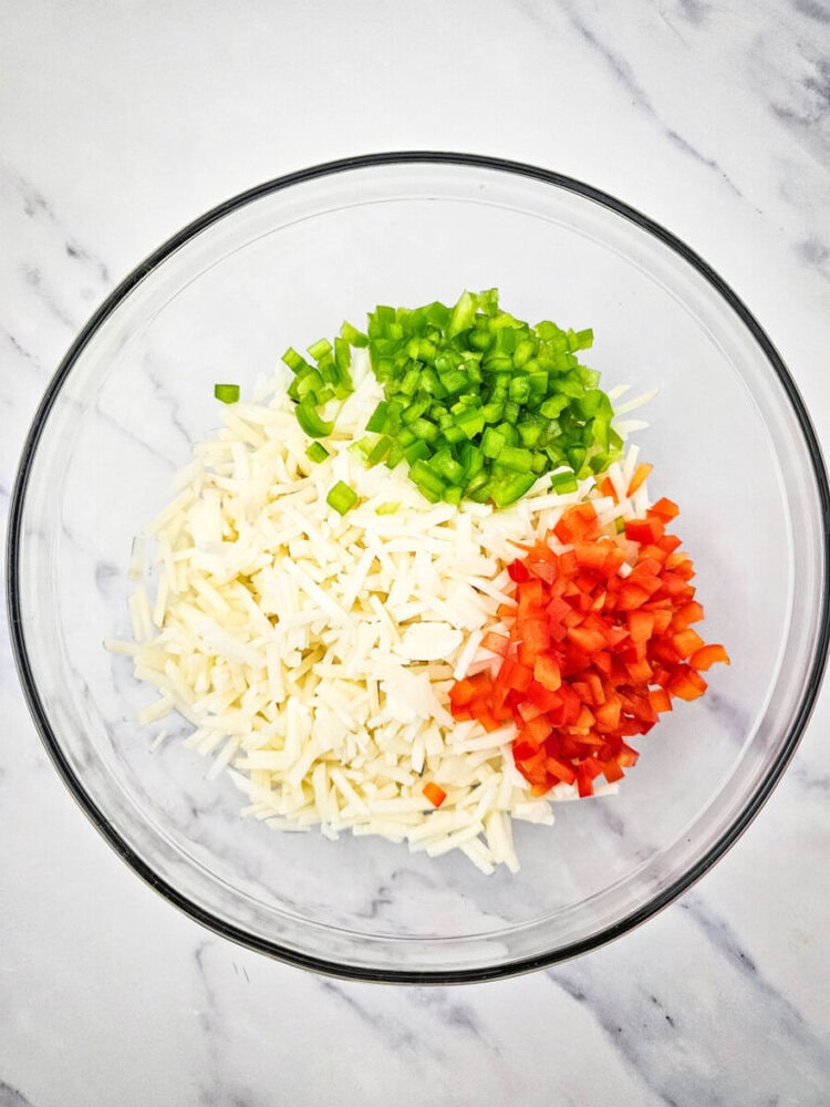 Glass bowl with chopped green bell pepper, red bell pepper, and shredded white potatoes on a marble surface.