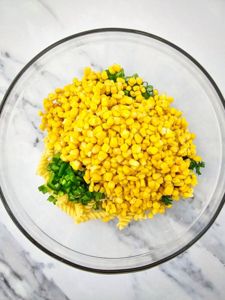 A glass bowl containing fusilli pasta, yellow corn, and chopped green onions on a marble surface.