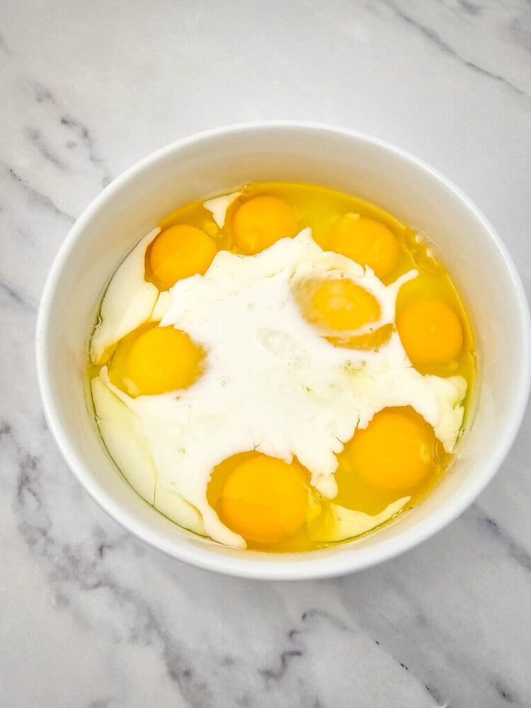 A white bowl containing six raw eggs, with clear egg whites and intact yolks, on a marble surface.