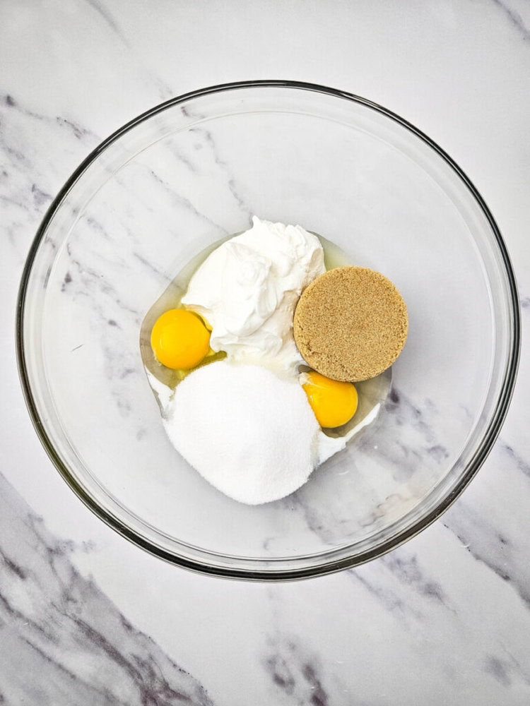 A glass bowl with ingredients including eggs, white sugar, brown sugar, and sour cream, placed on a marbled surface.