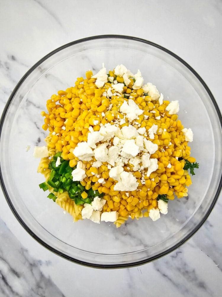 Glass bowl with cooked pasta, corn, chopped green onions, and crumbled white cheese on a marble surface.