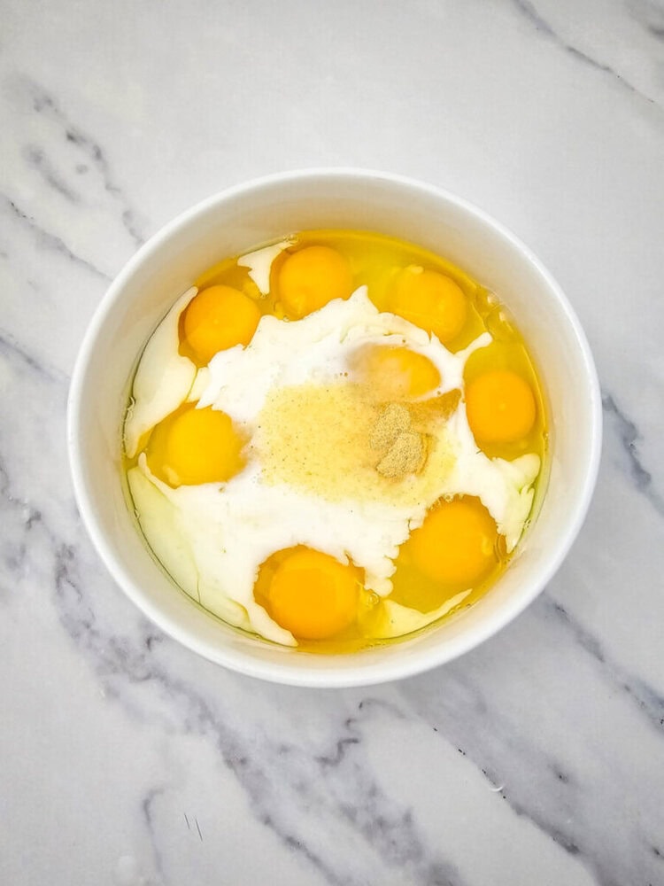 A bowl containing six raw eggs, milk, and a sprinkle of spices sits on a marble countertop.