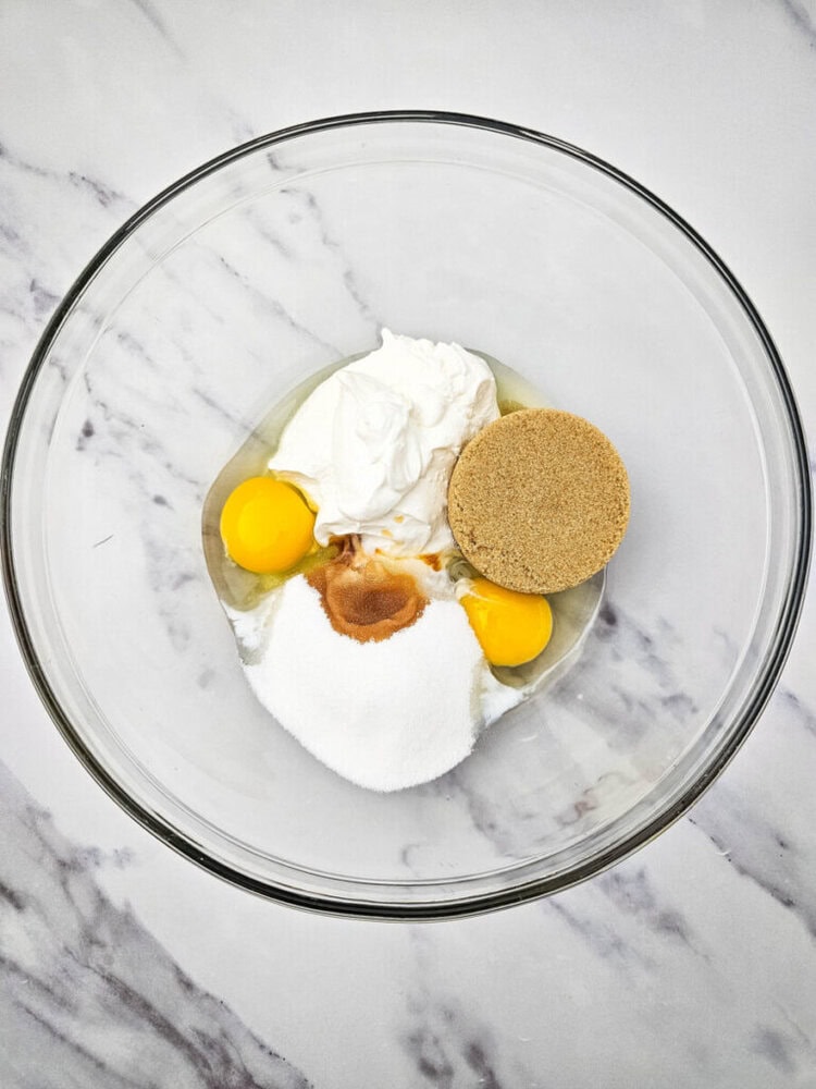 A glass bowl contains three eggs, a cup of yogurt, brown sugar, white sugar, and vanilla extract, on a marble countertop.