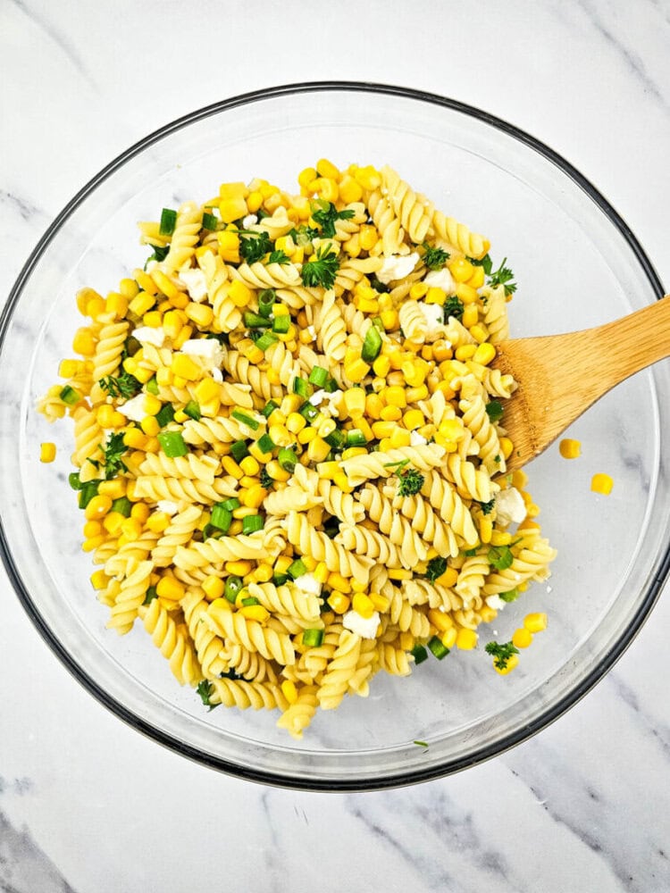 A glass bowl containing rotini pasta salad with corn, chopped green onions, and parsley, with a wooden spoon on the side.