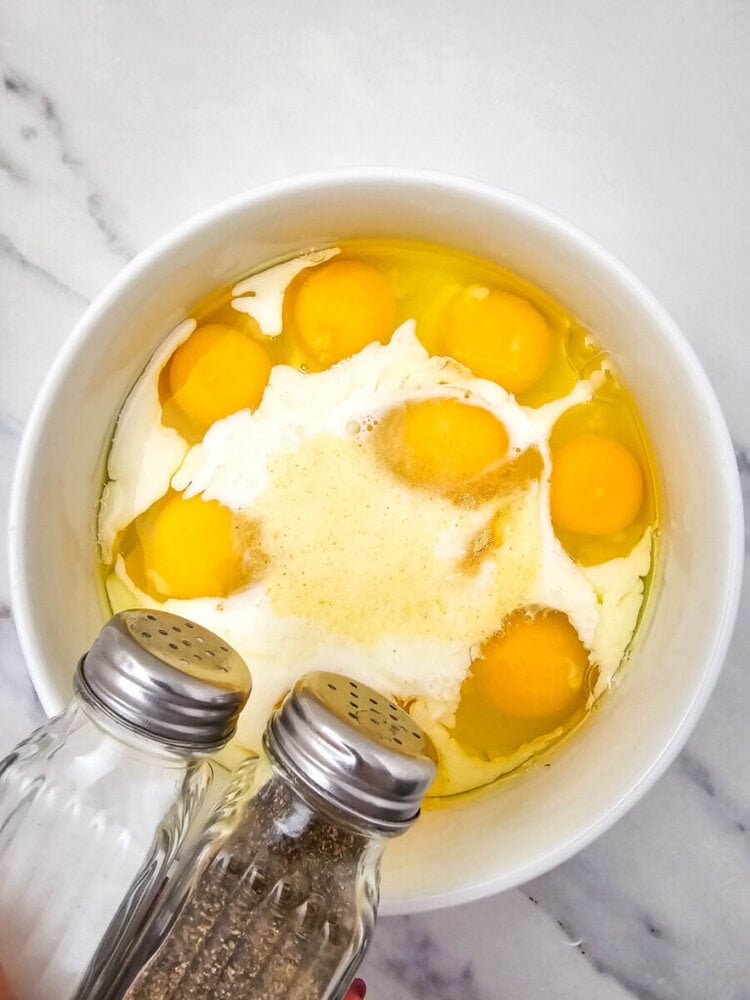 Bowl with raw eggs and milk, salt and pepper shakers positioned nearby, on a marble surface.