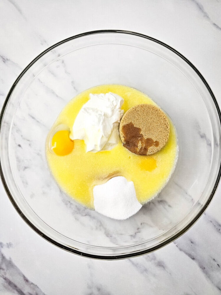Clear glass bowl with ingredients for baking: egg, sugar, brown sugar, melted butter, and sour cream on a marble countertop.