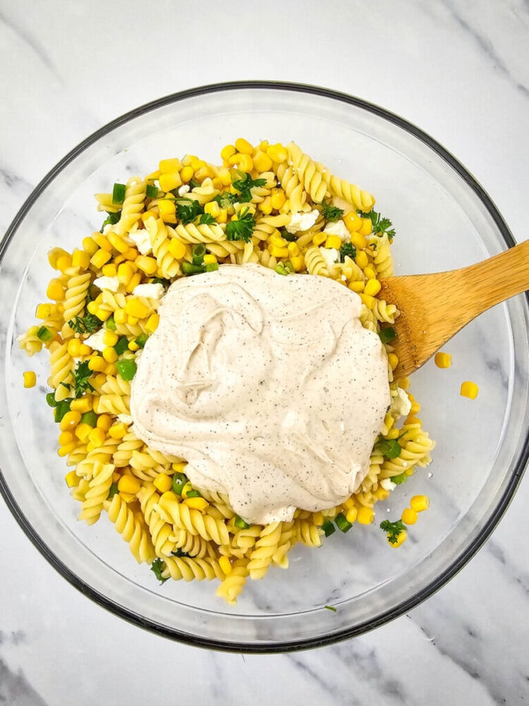 A glass bowl with pasta salad containing corn, peas, and parsley, topped with a dollop of creamy dressing. A wooden spoon is in the bowl, resting on a marble surface.