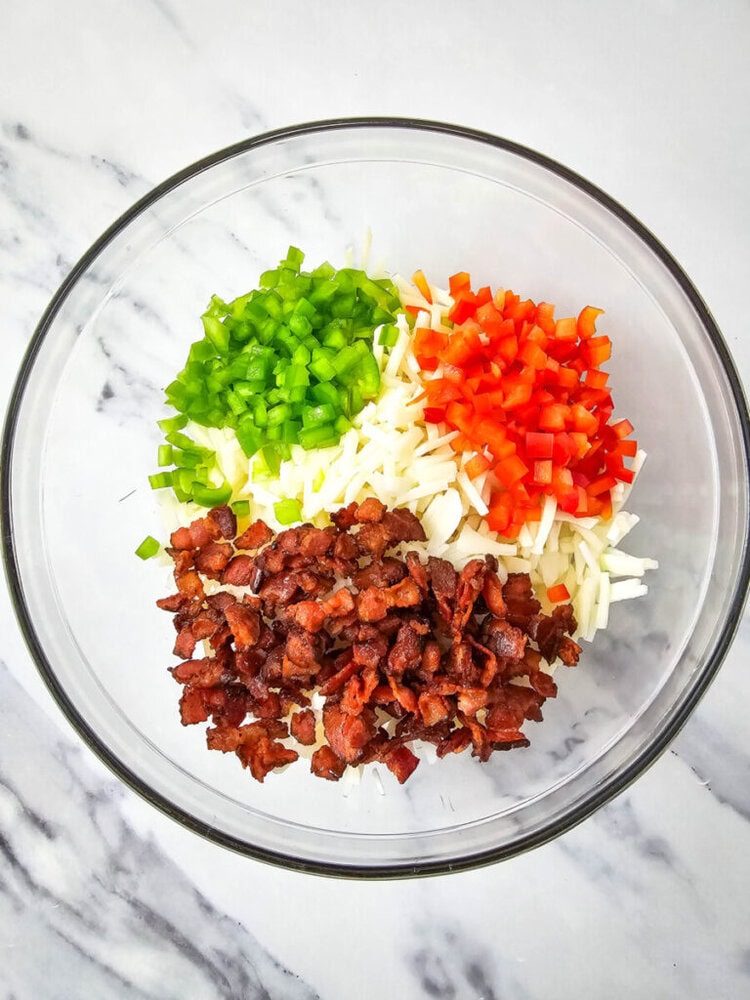 A glass bowl containing chopped green bell peppers, red bell peppers, shredded cheese, and cooked bacon pieces on a marble surface.