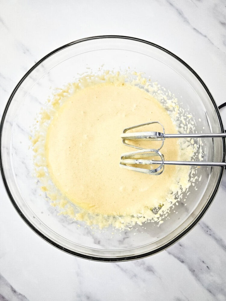 A clear glass bowl filled with beaten yellow batter. An electric mixer with two beaters is partially submerged in the mixture, sitting on a white marble surface.