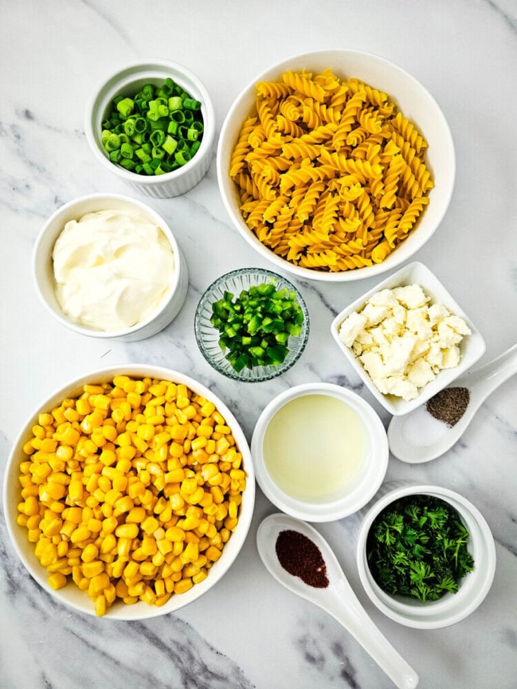 Bowls of ingredients on marble surface: rotini pasta, corn, mayonnaise, green onions, diced jalapeños, feta cheese, lime juice, black pepper, chili powder, and parsley.