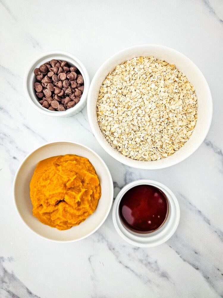 Bowls containing chocolate chips, oats, pumpkin puree, and a dark liquid placed on a white marble surface.