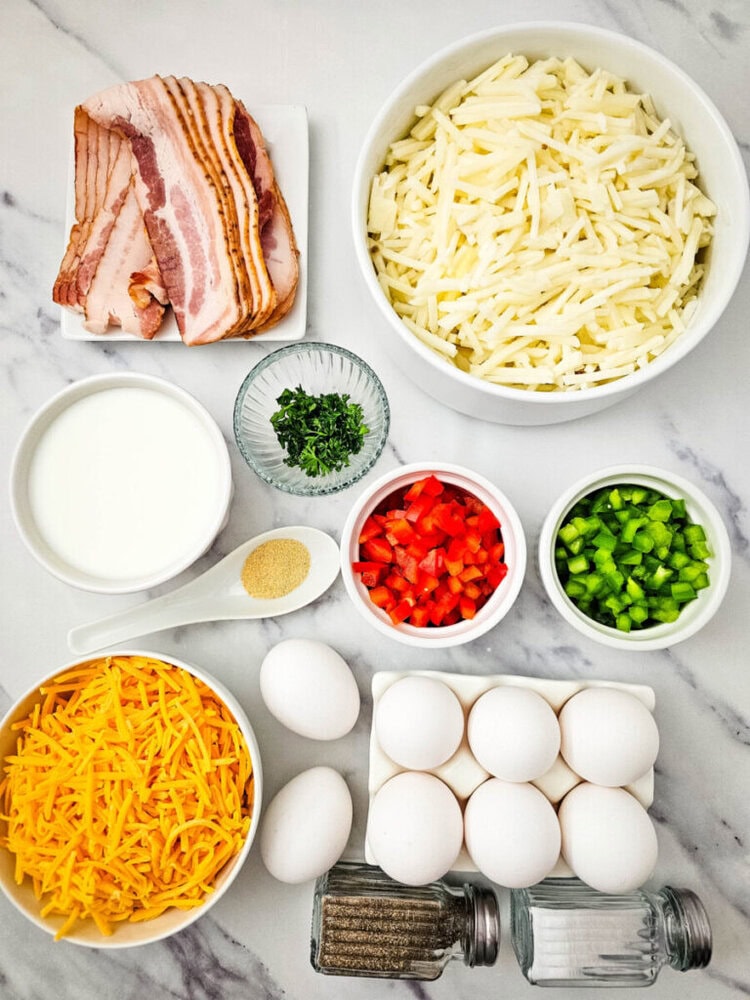 Ingredients on a marble surface include eggs, shredded cheese, milk, bacon, diced red and green bell peppers, shredded potatoes, seasoning, and chopped herbs.