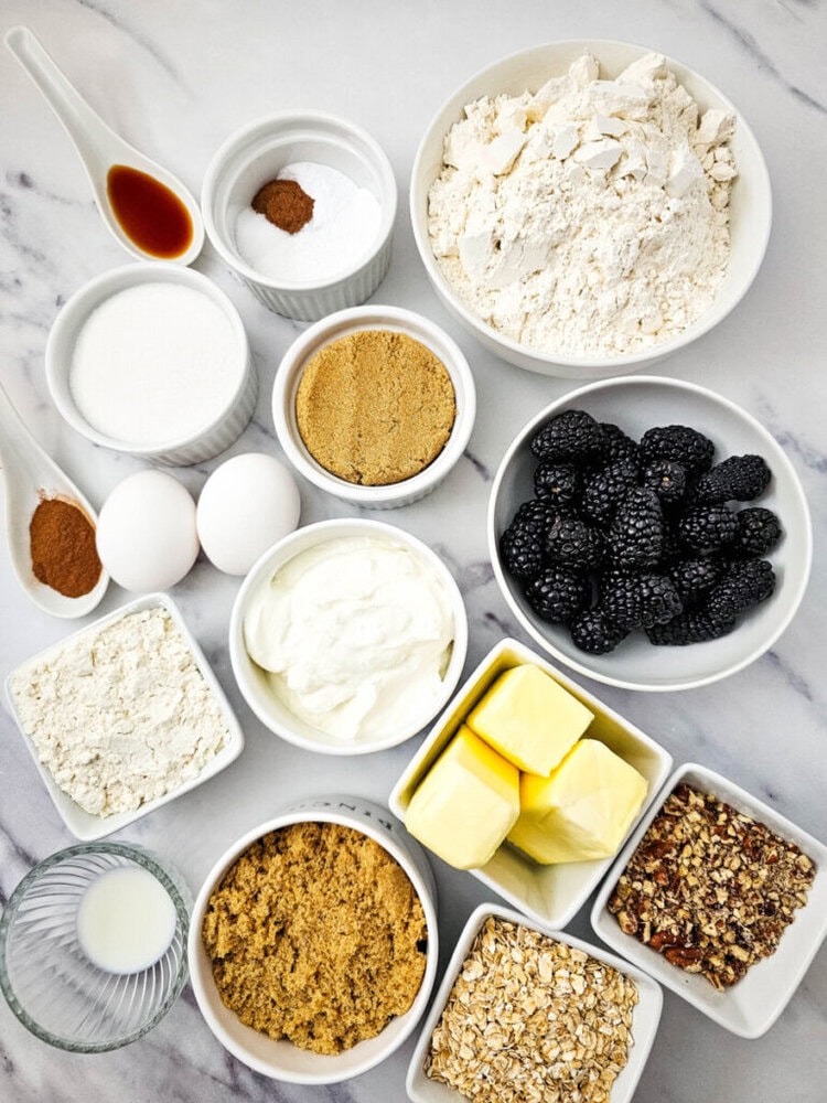 An assortment of baking ingredients is arranged, including flour, sugar, butter, yogurt, eggs, blackberries, oats, nuts, vanilla extract, and various spices on a marble surface.