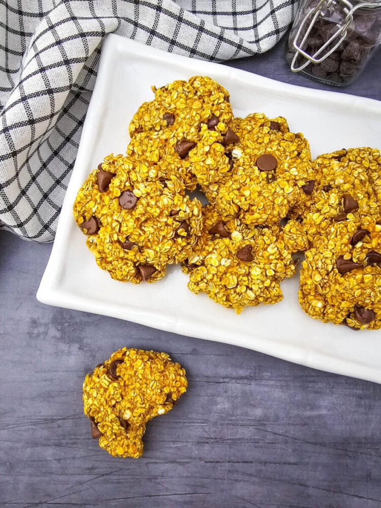 A plate of golden brown oat cookies with chocolate chips, one cookie partially eaten, sits on a gray surface next to a checkered cloth.