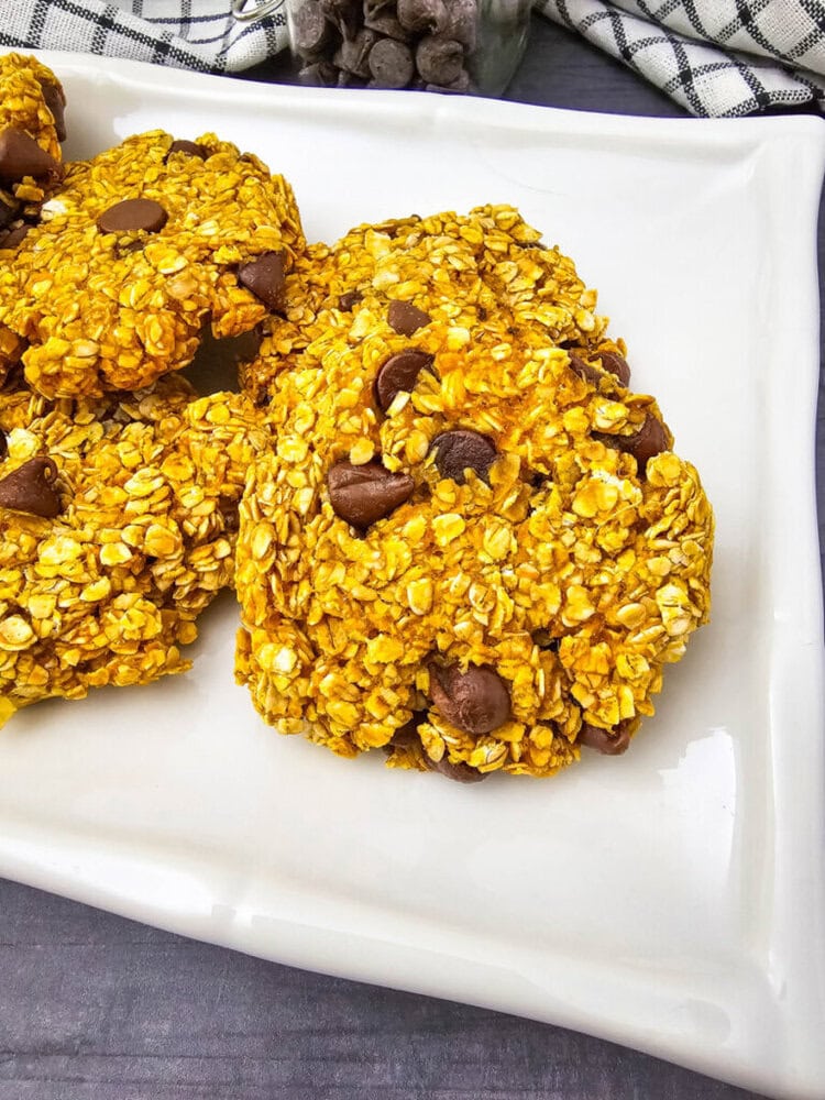 A white plate with cookies made of oats and chocolate chips, set on a dark surface with a checkered cloth peeking in the background.