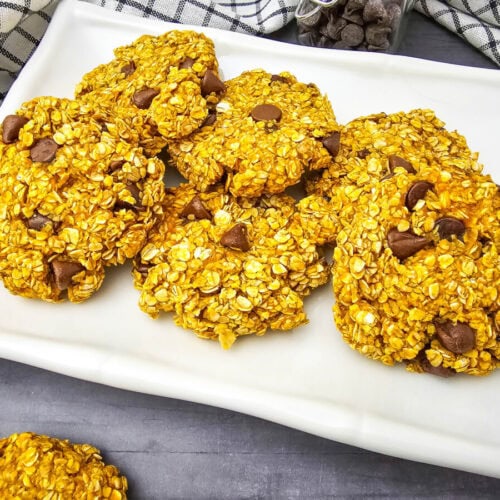 Plate of oatmeal cookies with chocolate chips, arranged on a white rectangular platter. Gray surface, checkered cloth, and small jar of chocolate chips in the background.