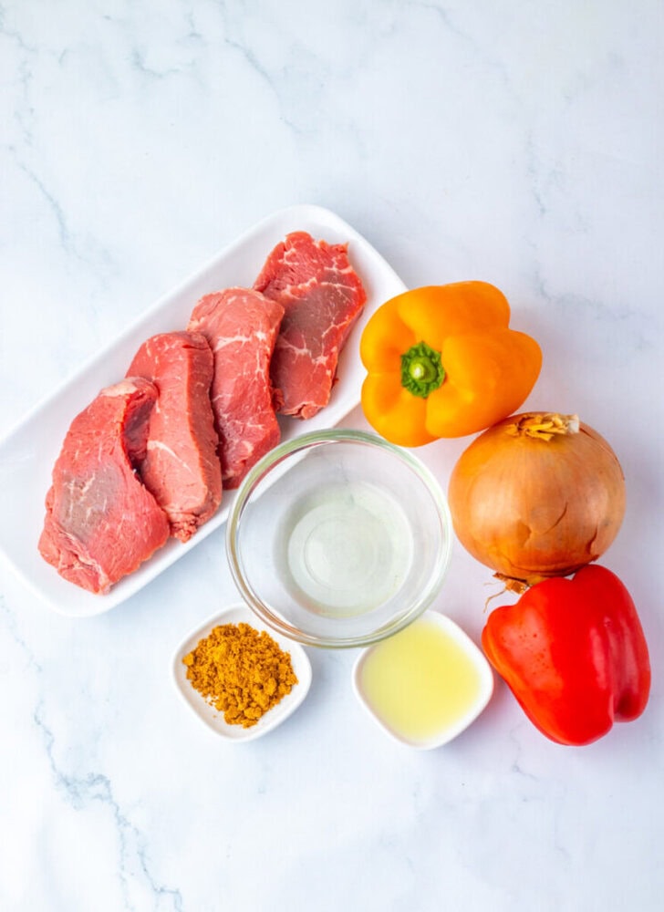 Three raw beef steaks on a white plate, a yellow bell pepper, a brown onion, a red bell pepper, a bowl of clear liquid, a small bowl of yellow seasoning, and a small bowl of oil.