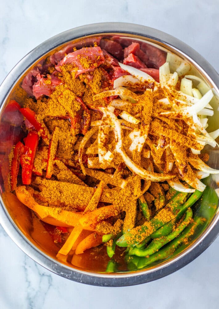 A metal bowl containing raw sliced beef, red, yellow, and green bell peppers, onion strips, and a generous amount of seasoning.
