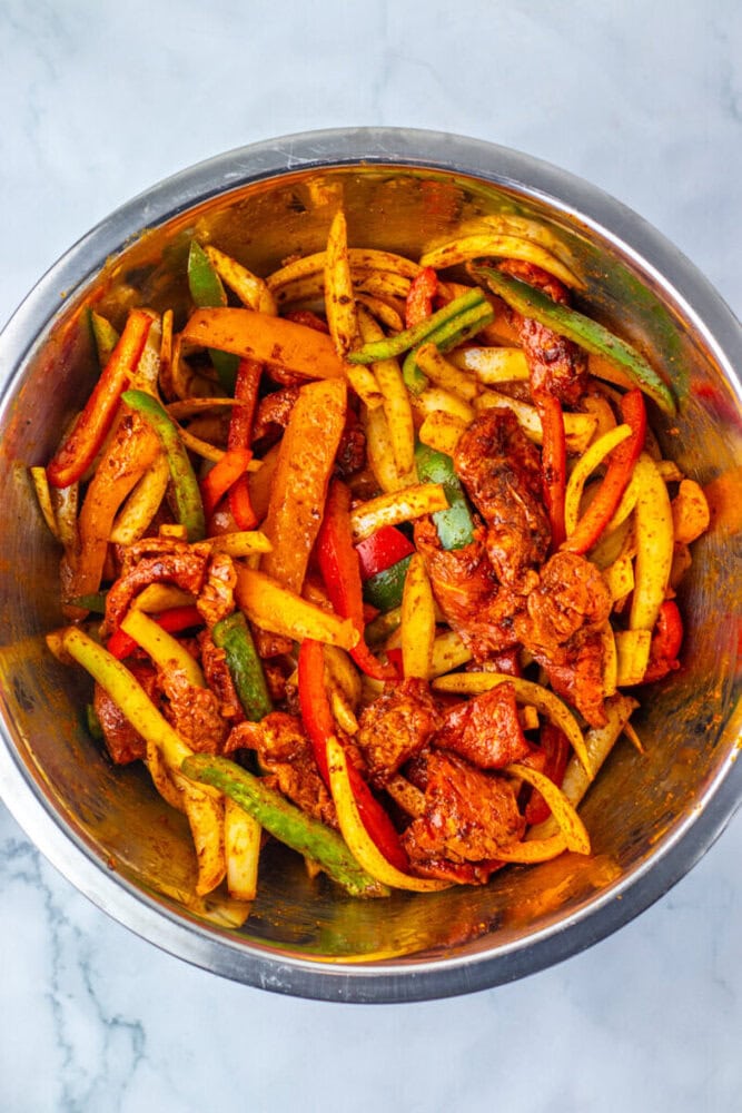 A metal bowl filled with sliced bell peppers, onions, and seasoned pieces of raw meat, mixed with spices and ready to be cooked.