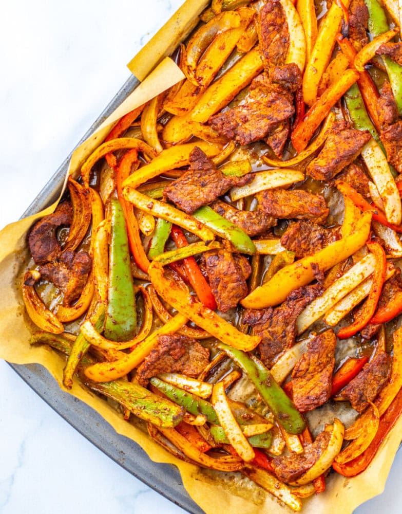 Sheet pan with cooked seasoned beef strips, yellow and green bell peppers, and onions on parchment paper, viewed from above.