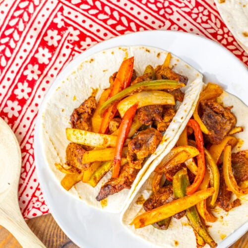 Two flour tortillas filled with seasoned beef strips and sautéed red and green bell peppers on a white plate, placed on a red and white patterned cloth.