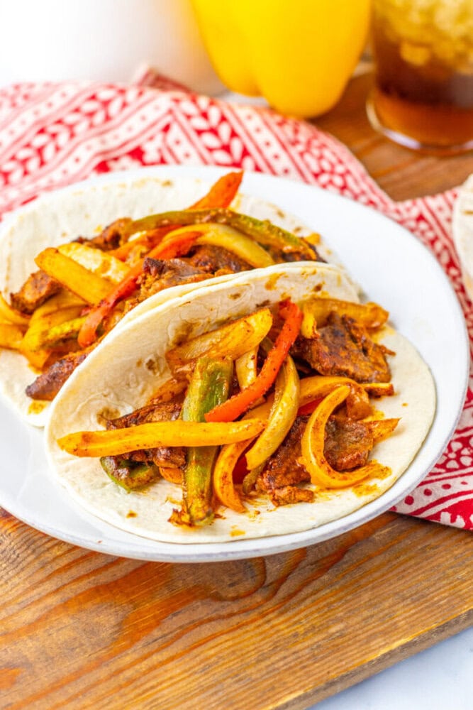 Two soft tortillas filled with seasoned beef strips, sautéed bell peppers, and onions on a white plate.