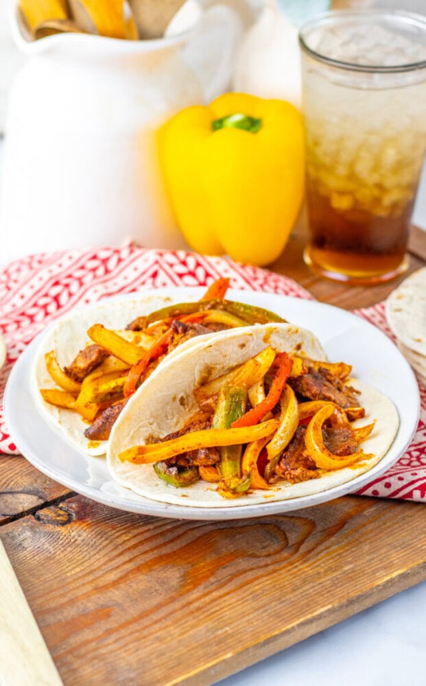 A plate with two fajitas filled with beef, onions, and bell peppers sits on a wooden tray next to a yellow bell pepper and a glass of iced drink.